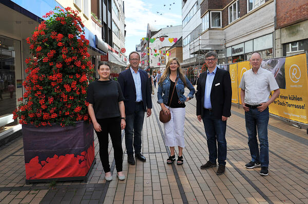 Die fünf Beteiligten stehen nebeneinander neben einer roten Topfpflanze in der Einkaufsstraße.
